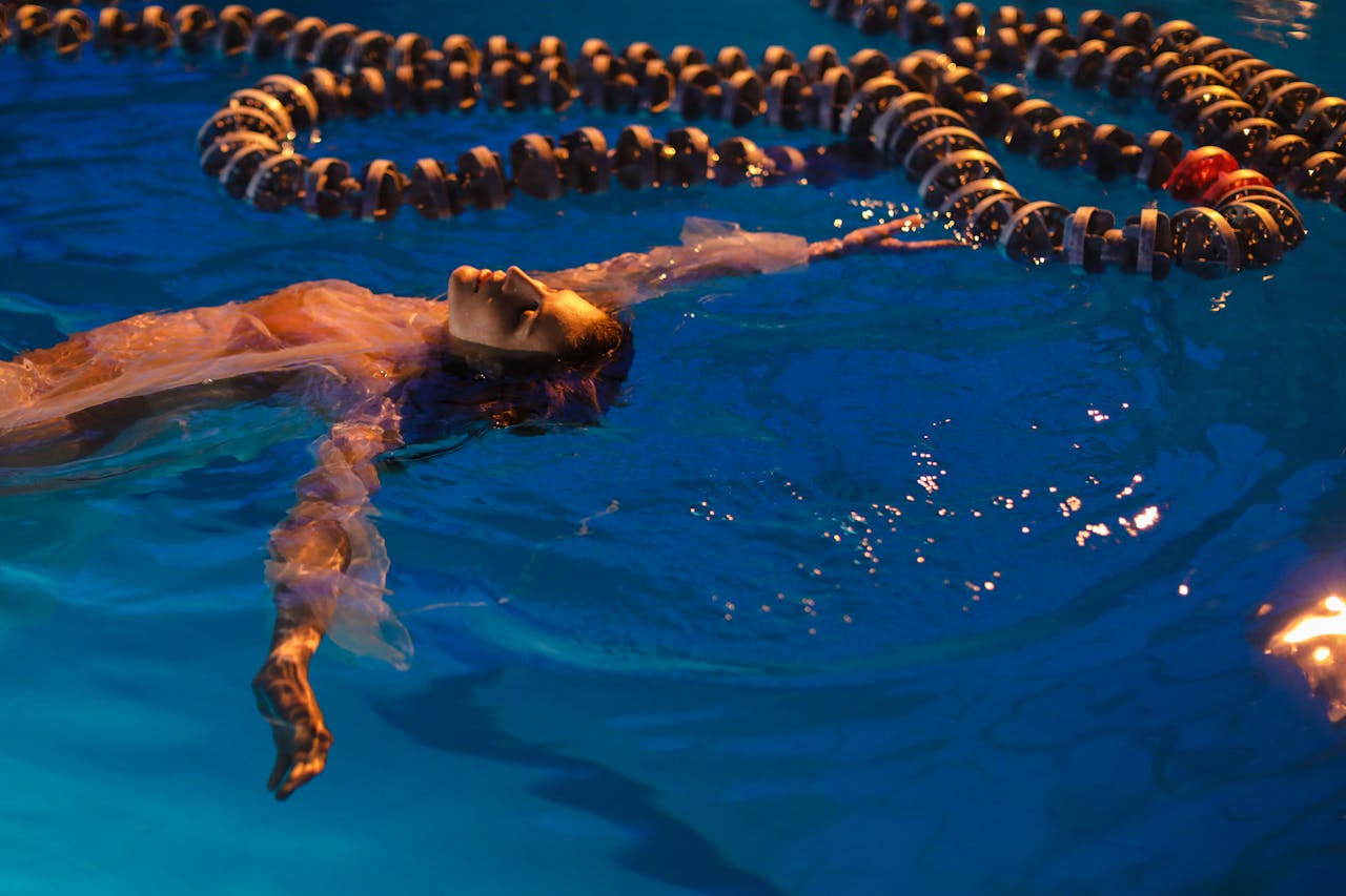 A woman floats calmly in a swimming pool under evening lights, surrounded by lane dividers.